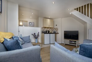 A modern living room with a grey sofa set, a flat-screen TV, and a kitchen area with a built-in oven and microwave. - Old Mill Court is a charming, dog-friendly two-bedroom cottage in Brixham (Brixham)