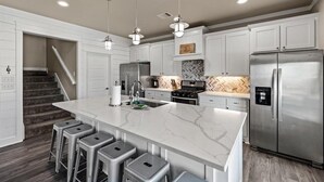 Een moderne keuken met witte kasten, een groot eiland met barkrukken, roestvrijstalen apparatuur en een backsplash met een herringbone-patroon.