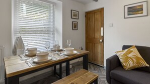 A dining area with a wooden table set for four, a bench, a sofa with a decorative pillow, and a door with a wooden panel. - Pisces Cottage, a delightful Grade II listed two bedroom cottage in Brixham (Brixham)