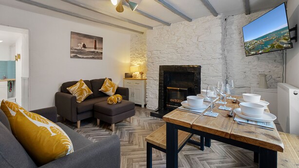 A modern living room with a fireplace, a dining table set for four, and a television mounted on the wall. - Pisces Cottage, a delightful Grade II listed two bedroom cottage in Brixham (Brixham)