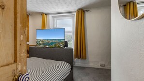 A bedroom with a bed, a television displaying a scenic view, a window with curtains, and a door. - Pisces Cottage, a delightful Grade II listed two bedroom cottage in Brixham (Brixham)
