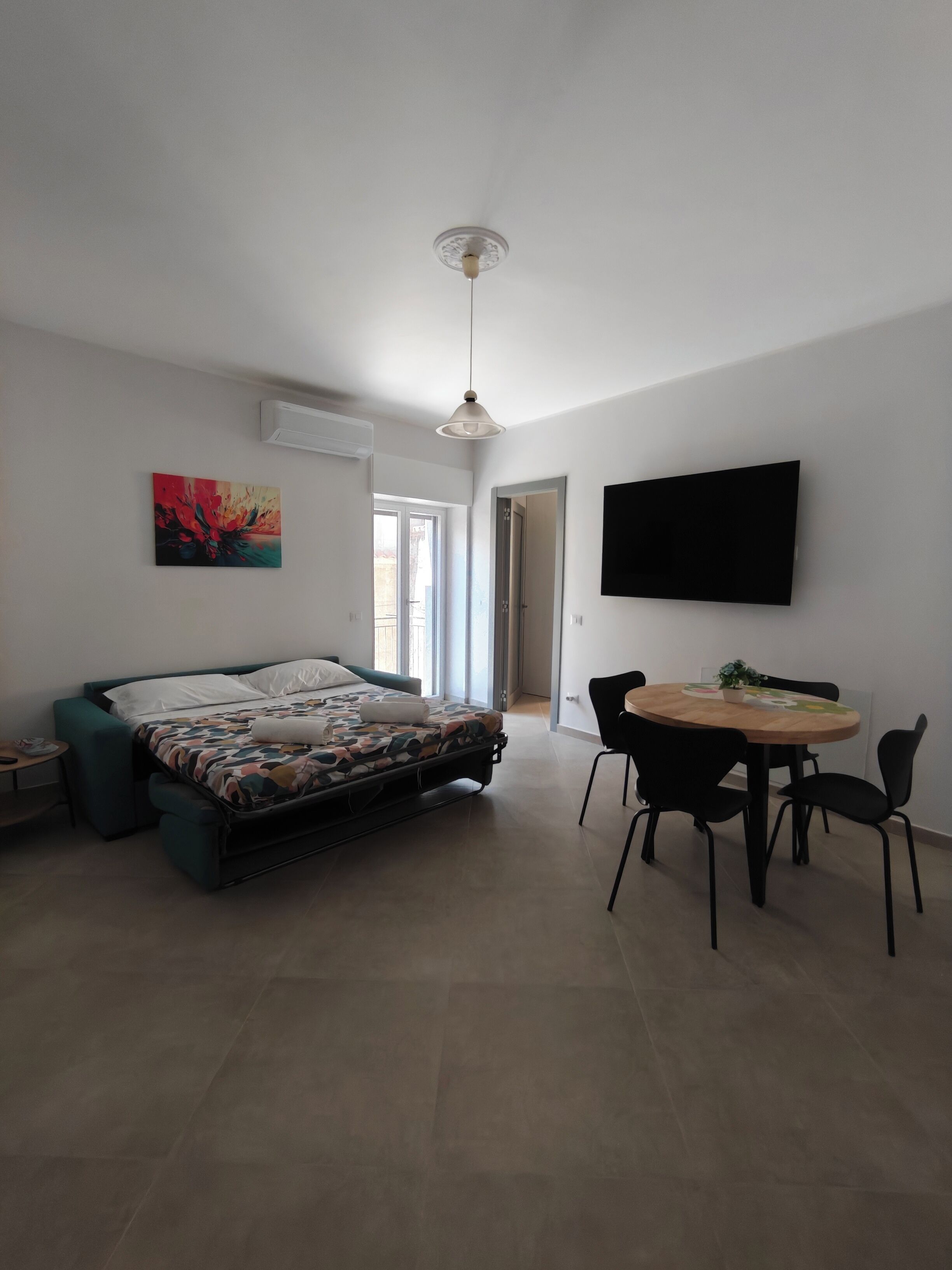 A modern bedroom with a bed, a dining table with chairs, a TV mounted on the wall, and a window.