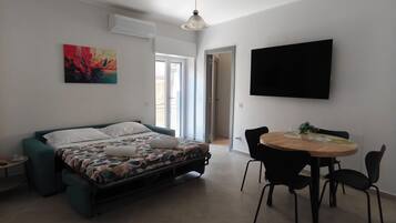 A modern bedroom with a bed, a dining table with chairs, a TV mounted on the wall, and a window.