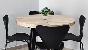 A round wooden table with four black chairs, a small potted plant, and a white wall in the background.