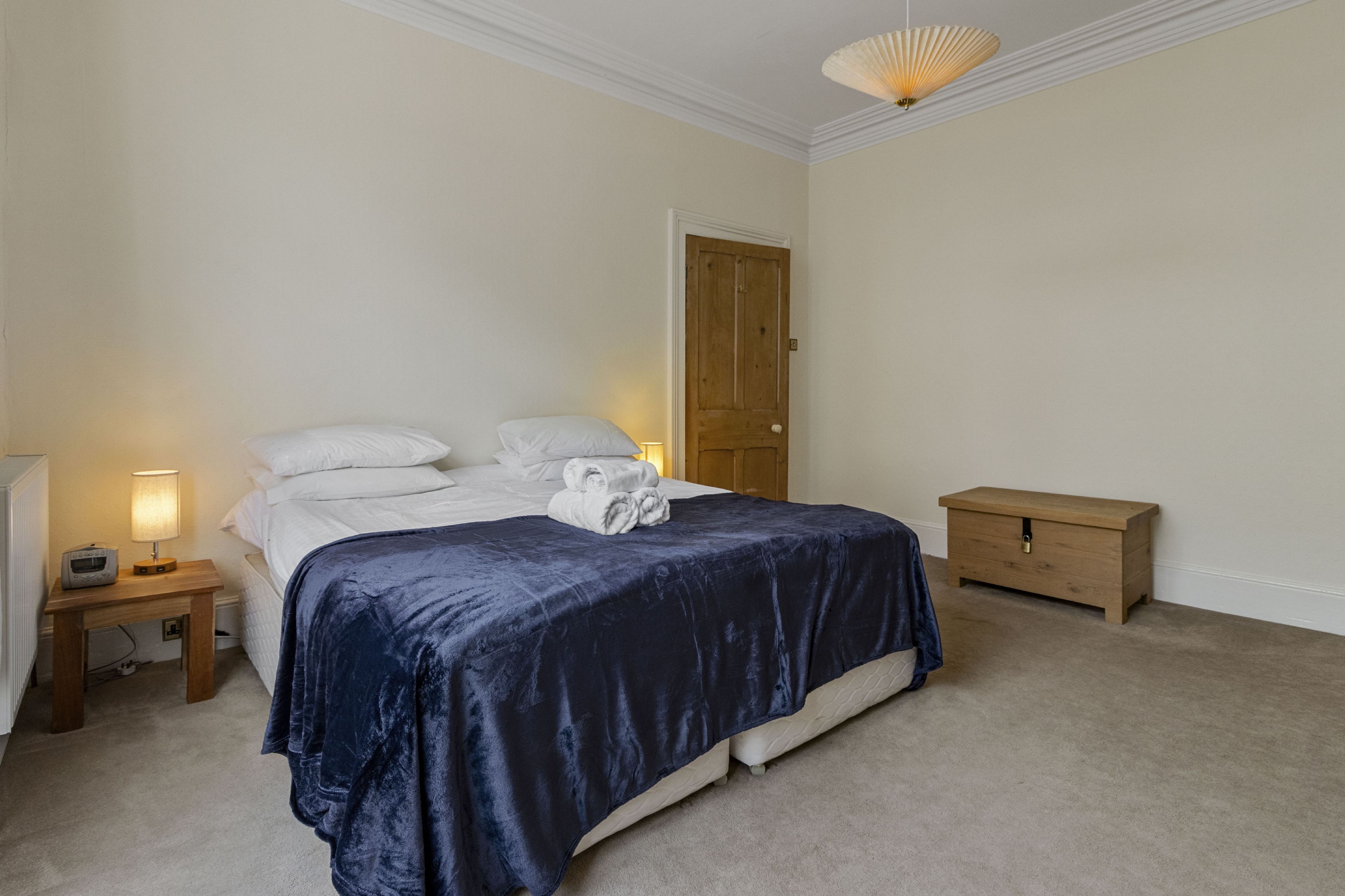 Uma cama bem arrumada com um cobre-leito azul escuro, uma mesa de cabeceira de madeira com um abajur e um aparador de madeira num quarto com paredes brancas e uma porta de madeira.