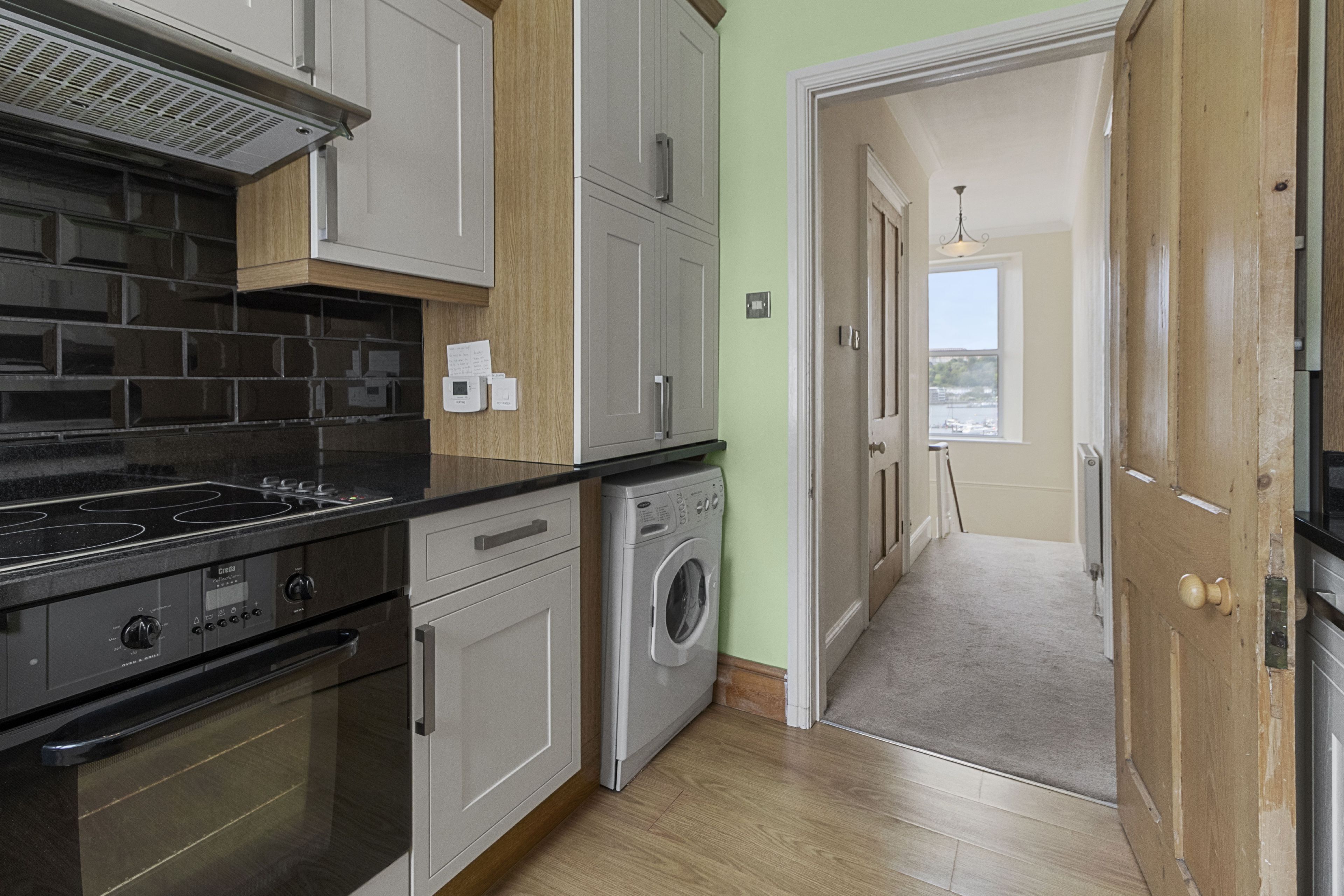 Une cuisine équipée d’une cuisinière, d’un four et d’un lave-linge, avec des meubles de rangement en bois et un couloir menant à une porte.