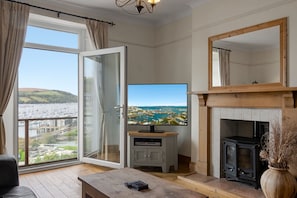 A living room with a TV, fireplace, and a view of a marina. - Marina Cottage - a charming Kingswear holiday cottage overlooking the River Dart (Kingswear)