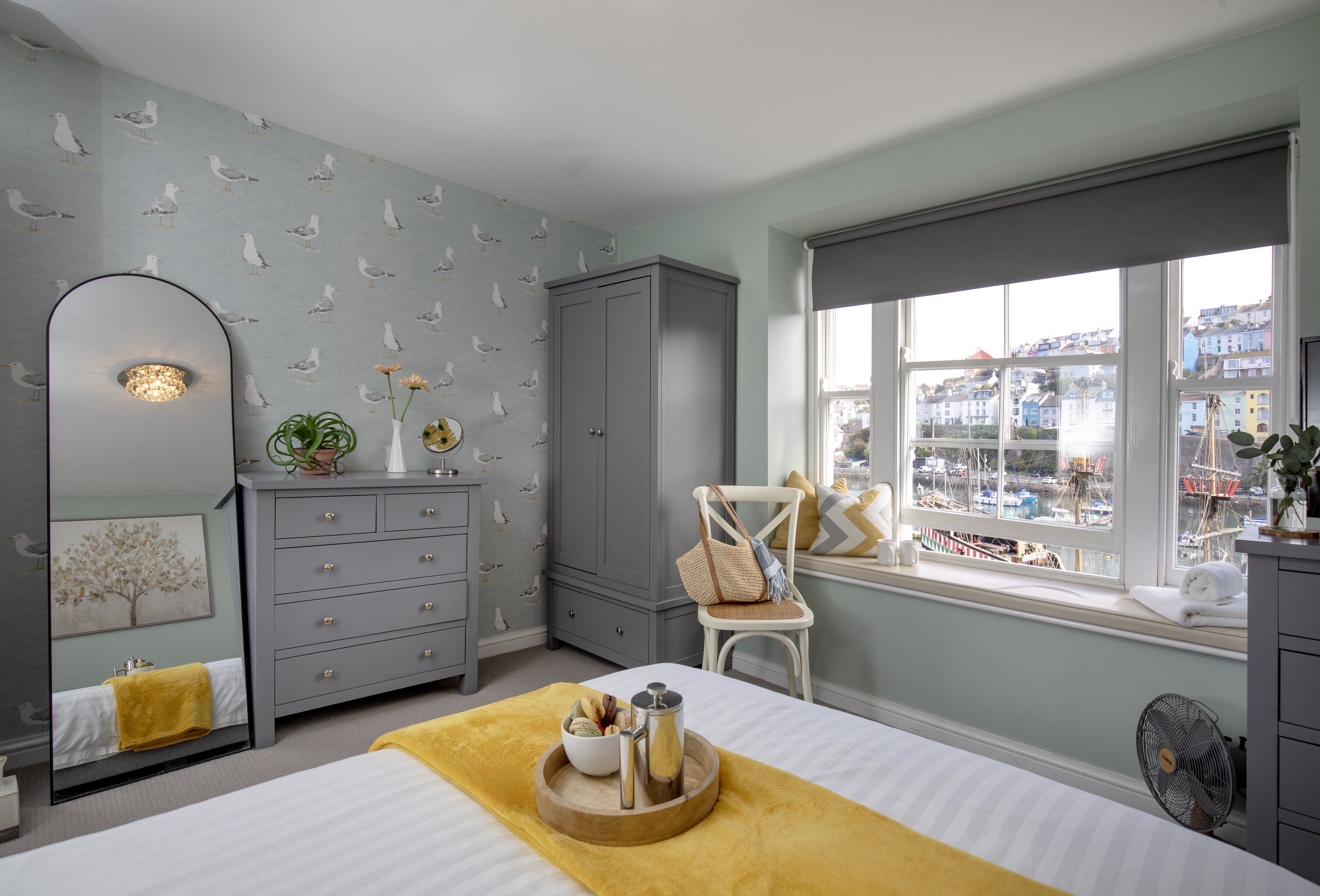 A bedroom with a bed, dresser, mirror, window with a view of boats, and a dresser with a mirror.