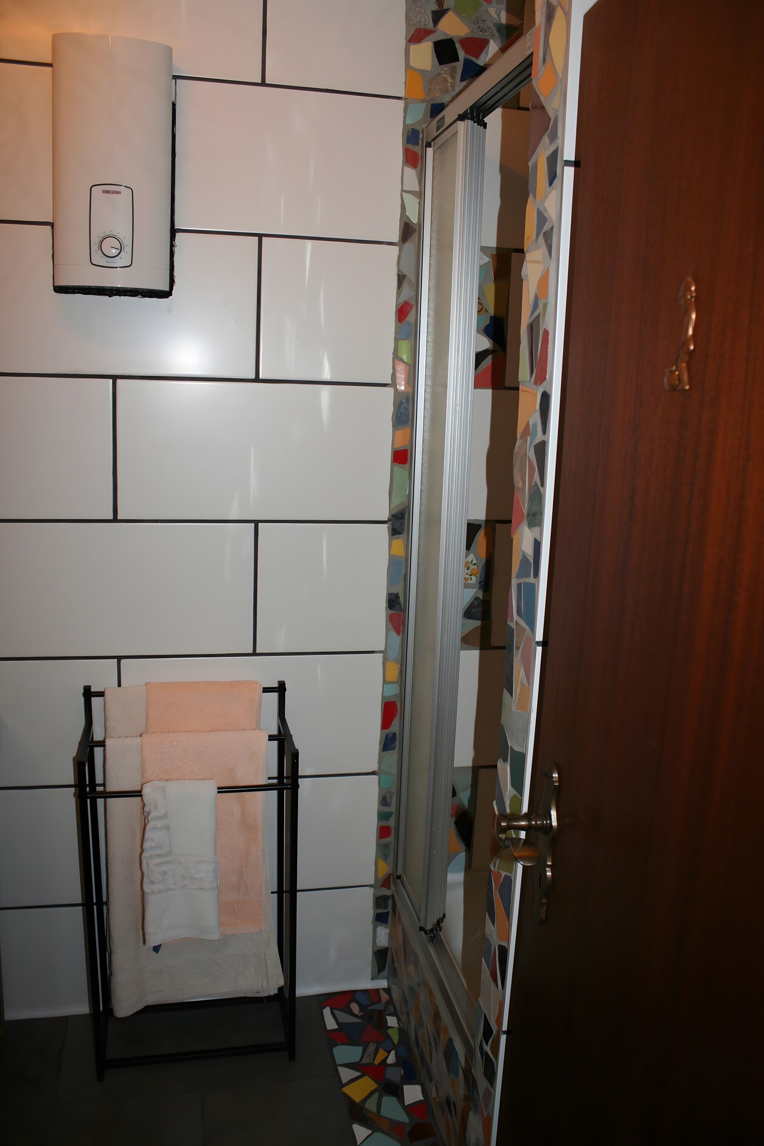 Un bagno con pareti piastrellate di bianco, un portasciugamani con un asciugamano bianco, una doccia con bordo in mosaico colorato e una porta in legno.