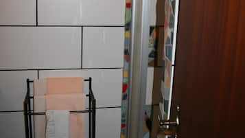 A bathroom with white tiled walls, a towel rack with a white towel, a shower with a colorful mosaic tile border, and a wooden door.