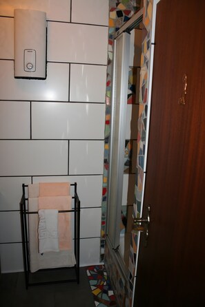 A bathroom with white tiled walls, a towel rack with a white towel, a shower with a colorful mosaic tile border, and a wooden door. - Apartment for 2 Guests (39 m²) in Ludwigswinkel (Dogs Welcome) (Ludwigswinkel)