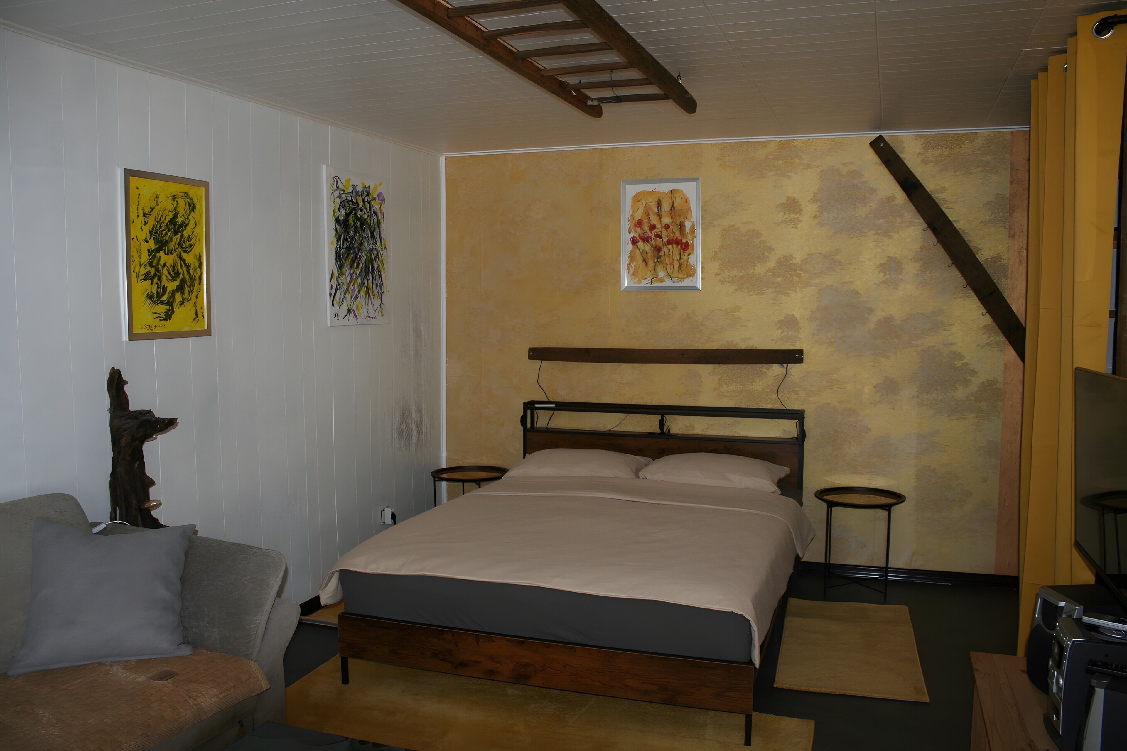 A bedroom with a bed, a chair, a wooden headboard, a ladder, and artwork on the walls.