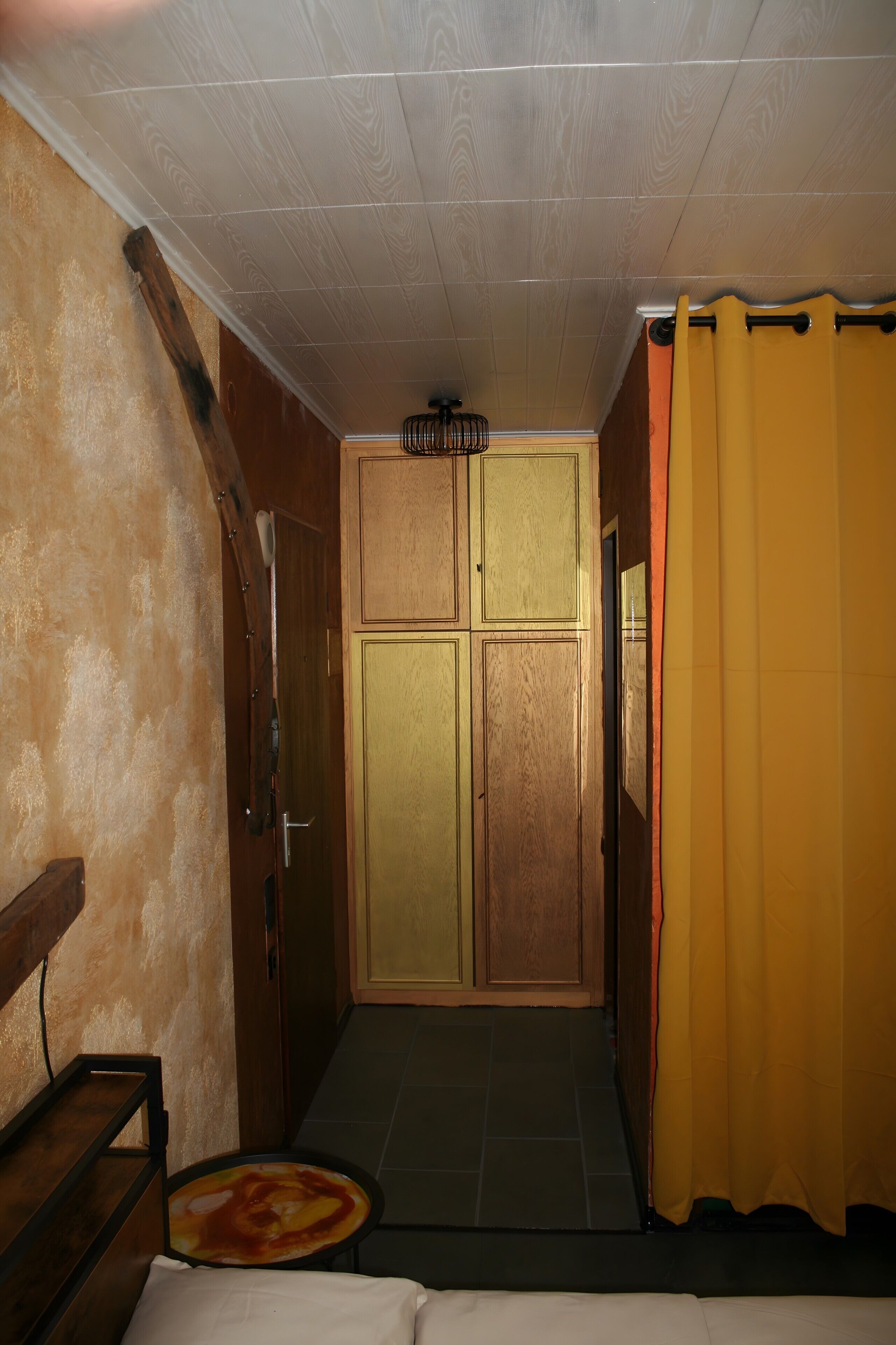 A narrow hallway with a wooden door, a small table with a pizza, and a yellow curtain.