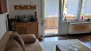 A living room with a sofa, wooden cabinets, a radiator, and a window with a view of a fence. - Apartment for 2 Guests (39 m²) in Ludwigswinkel (Pets Welcome) (Ludwigswinkel)