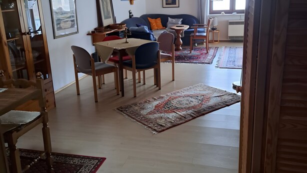 Olohuone, jossa on puinen katto, ruokailutila pöytineen ja tuoleineen, tyynyillä varustettu sohva ja suuri ikkuna, jossa on verhot.