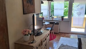 A room with a desk, a computer, a vase of flowers, and a window with a view of greenery.