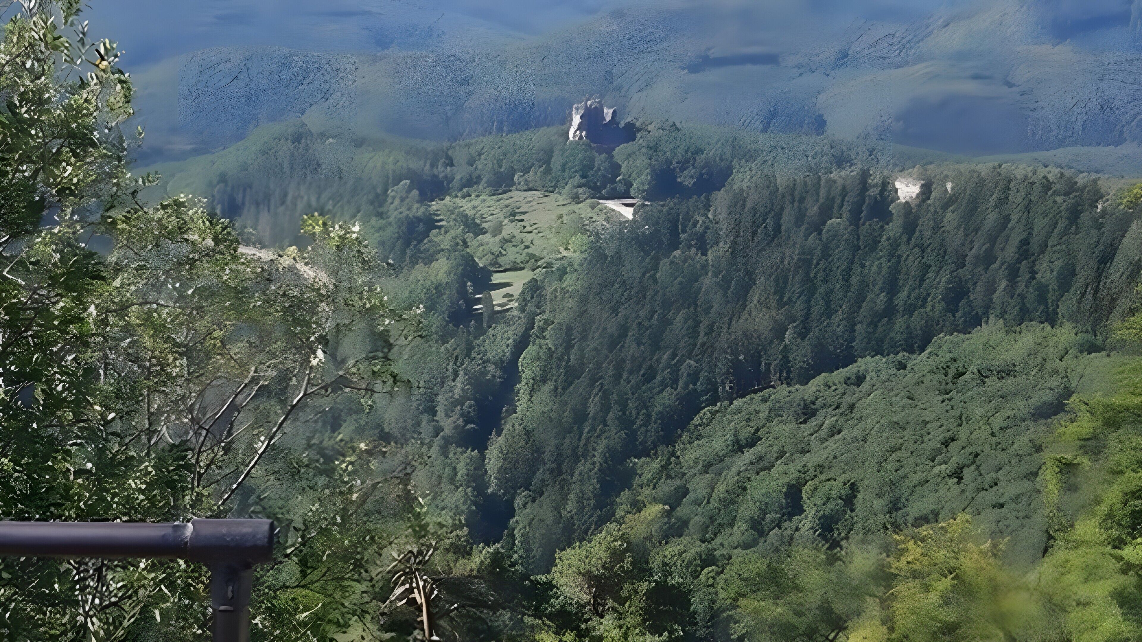A mountainous landscape with dense forest, a clear sky, and a distant rocky outcrop.