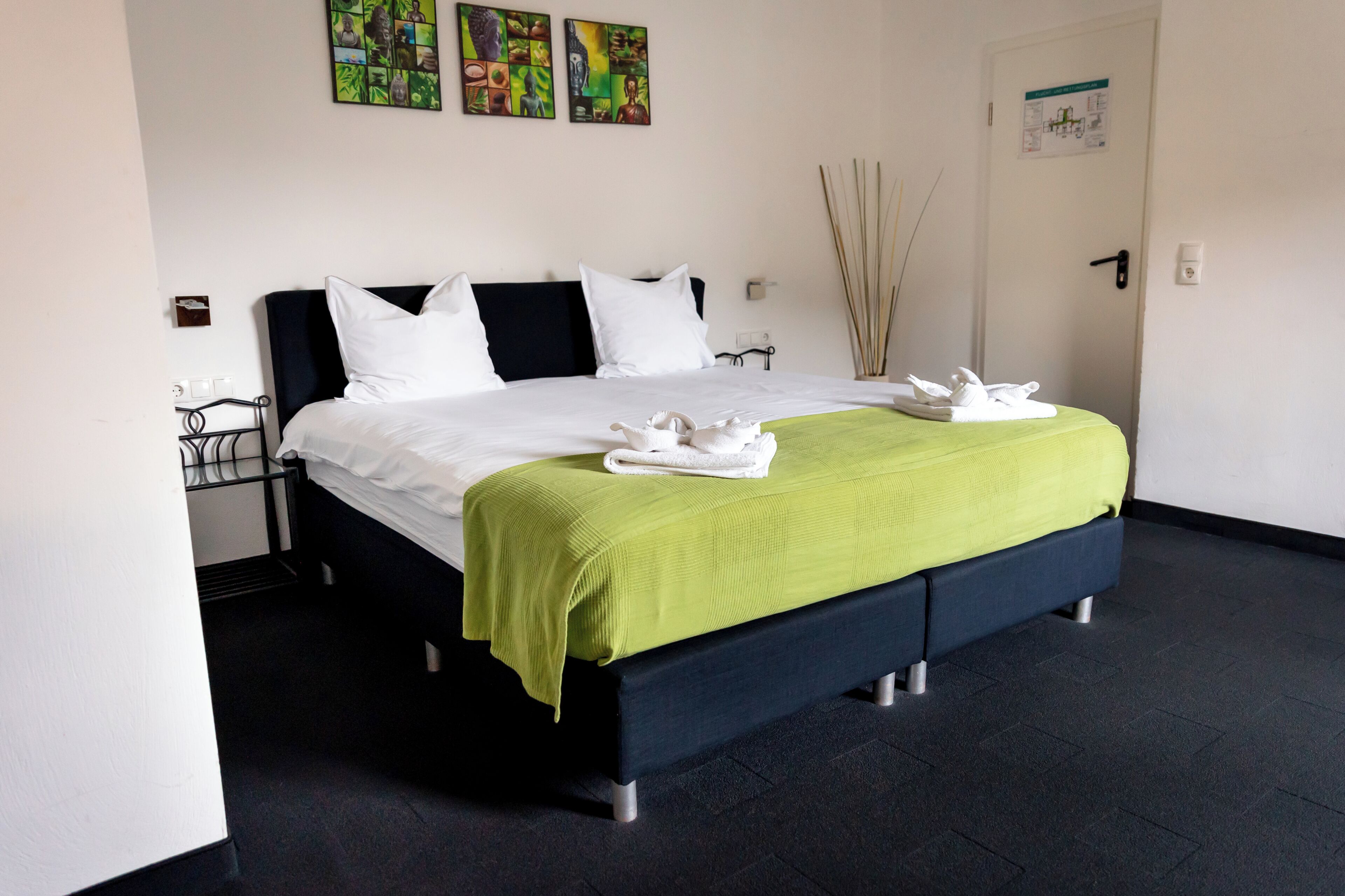 A neatly made bed with white linens and a green throw, a black headboard, and a wall-mounted artwork.