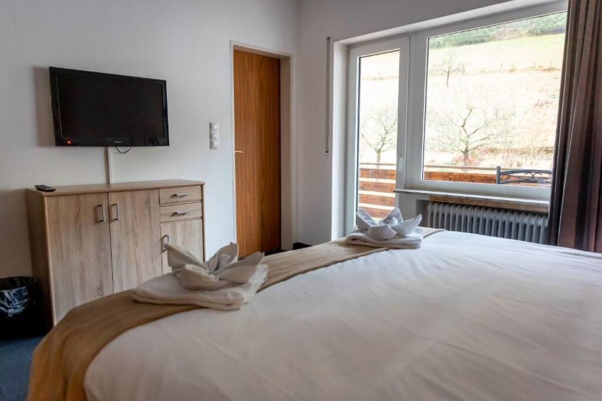 A hotel room with a bed, a flat-screen TV, a wooden wardrobe, and a window with a view of a field.