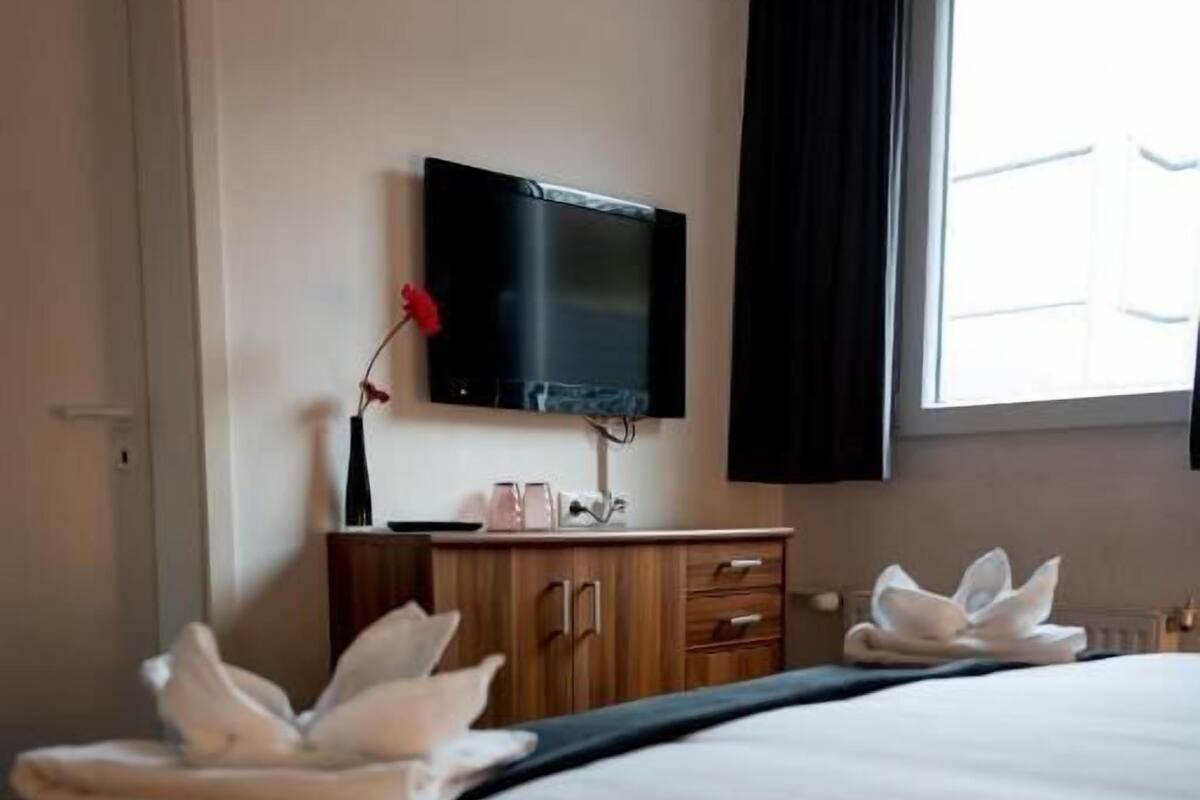 A hotel room with a bed, a TV mounted on the wall, a wooden dresser, and a window with curtains.