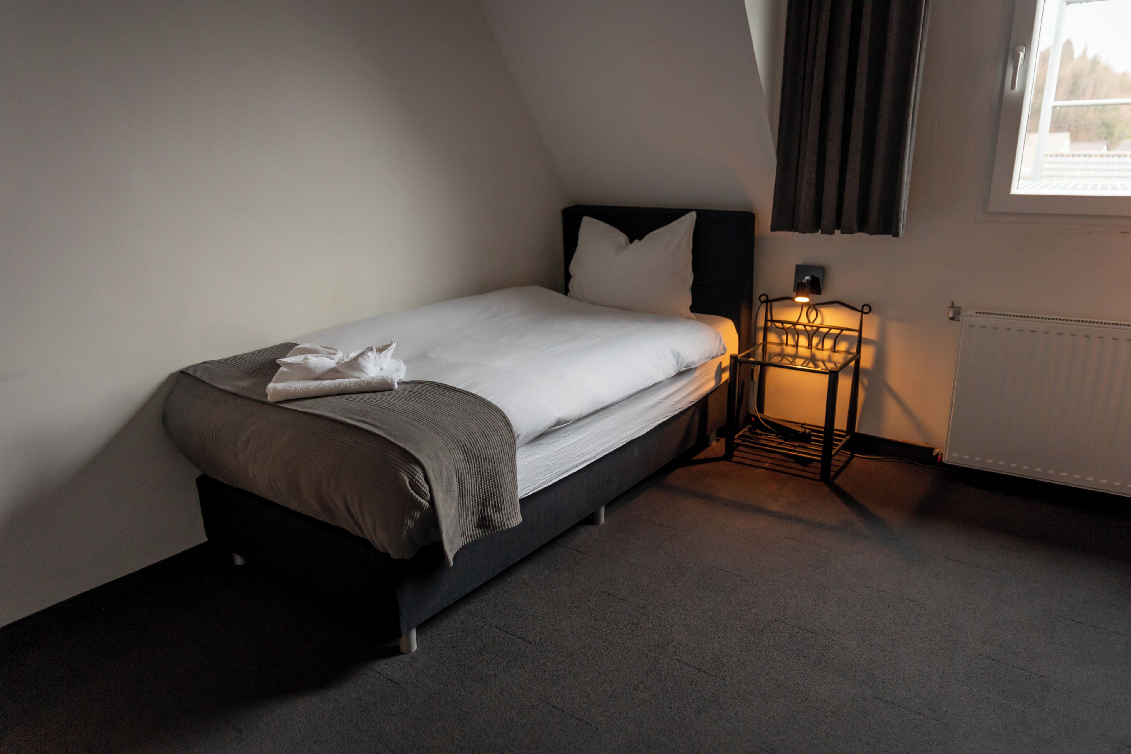 Een enkel bed met hoofdeinde, een nachtkastje met lamp en een raam met gordijnen.
