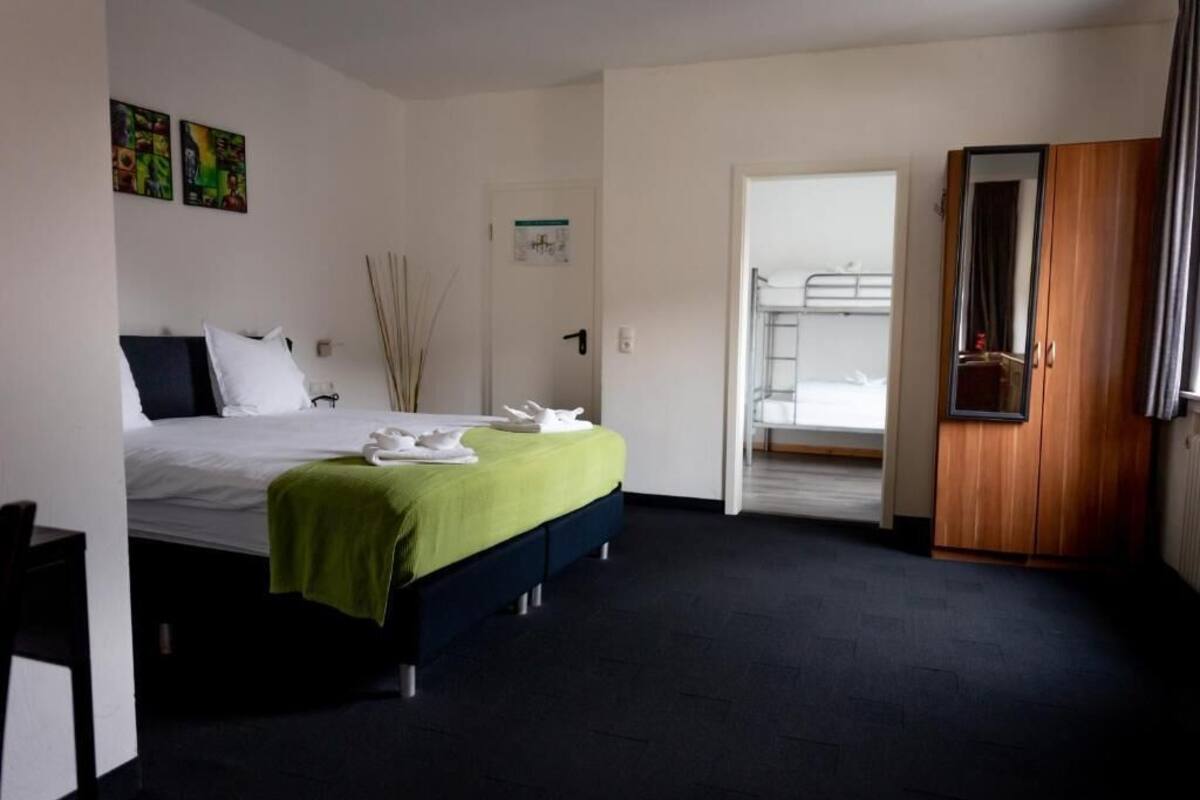 A hotel room with a bed, a wooden wardrobe, a door leading to a bathroom, and a small table.