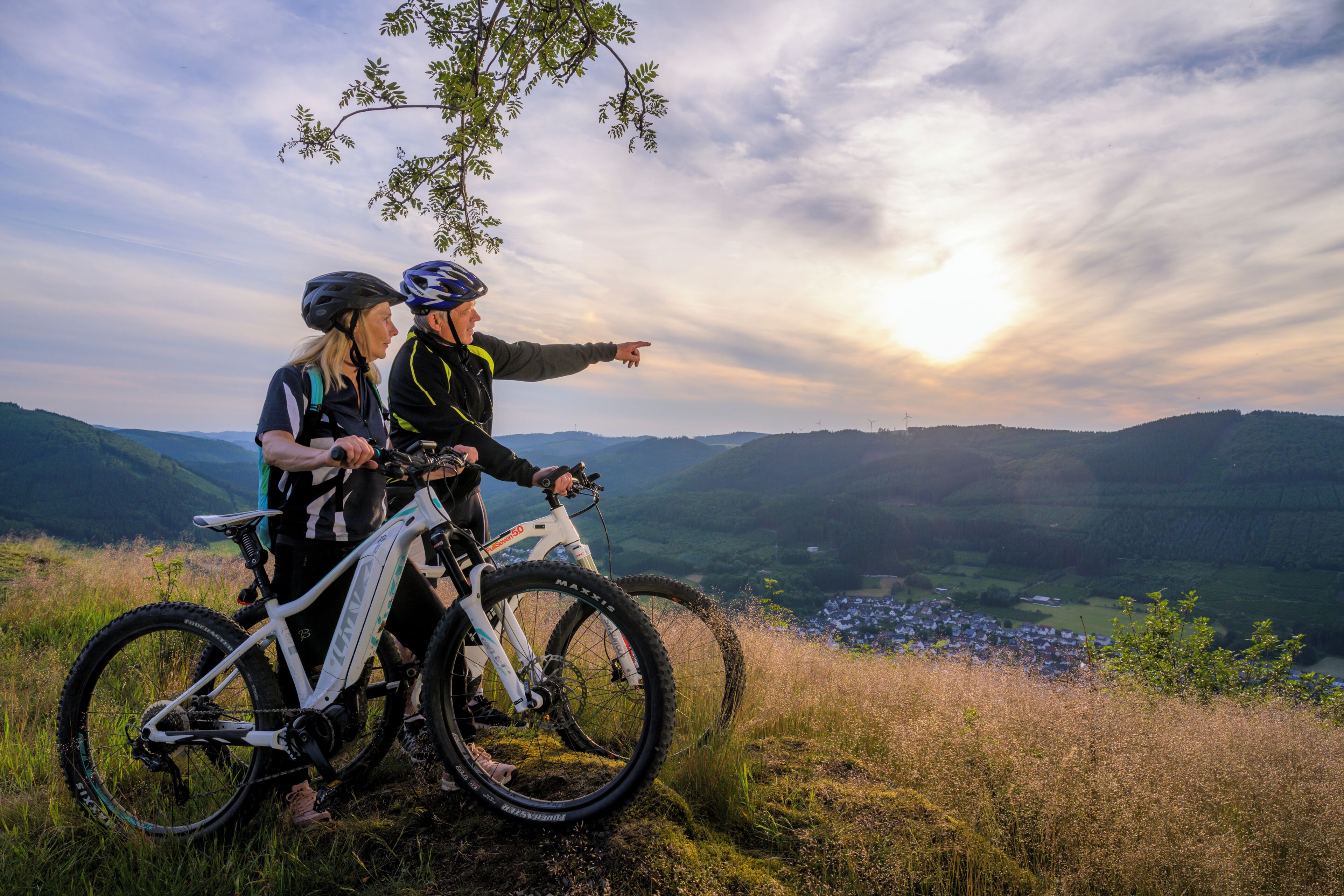 Dos personas en un sendero de bicicleta de montaña con una vista panorámica de un valle y un pueblo distante.