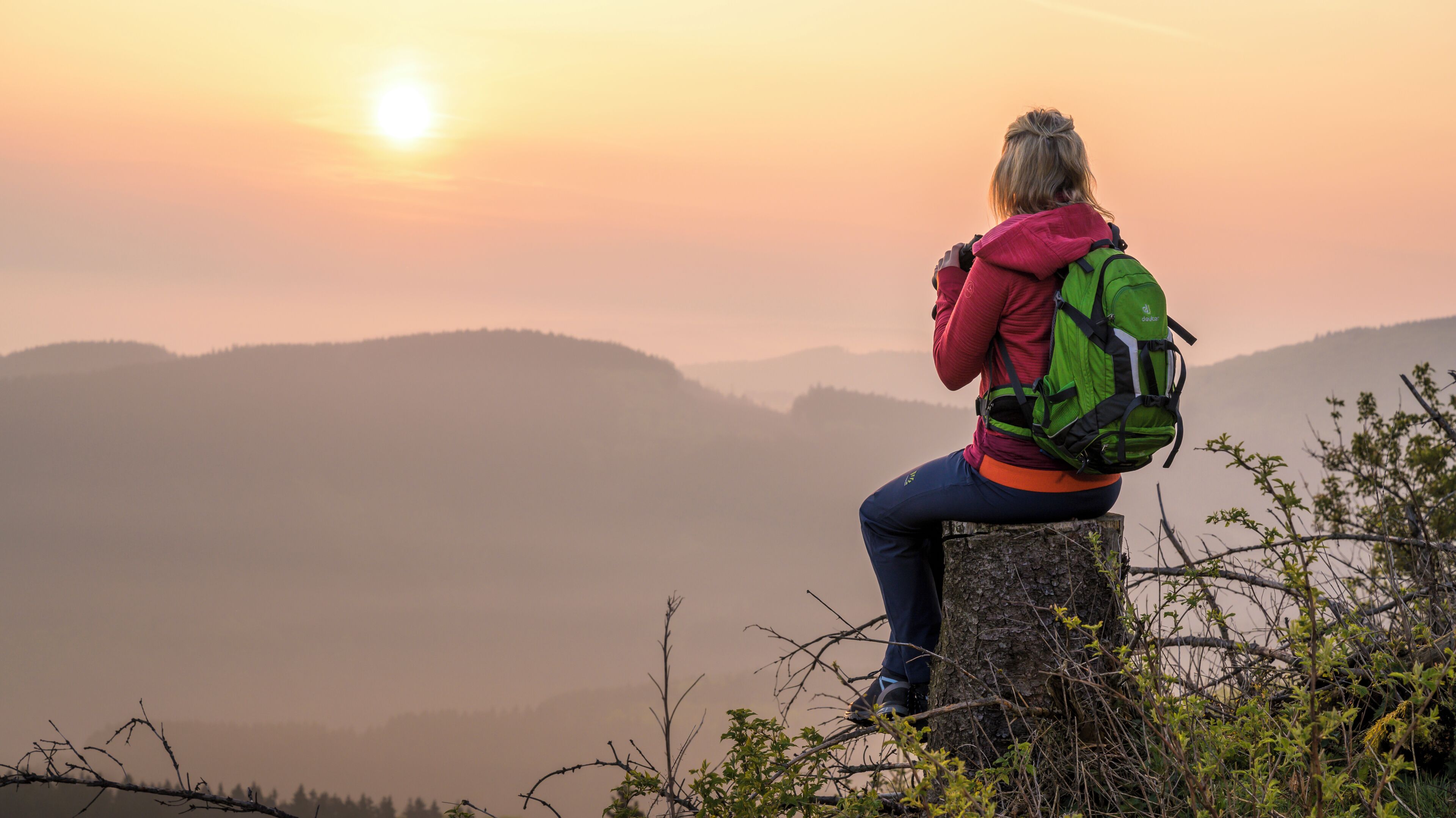 A person sitting on a rock with a green backpack, overlooking a mountainous landscape during sunset.