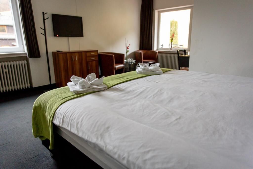 Ein Hotelzimmer mit einem großen Bett, einem flachbildfernseher an der Wand, einem Holzschrank, einer Sitzecke mit einem Sessel und einem kleinen Tisch sowie einem fenster mit Vorhängen.
