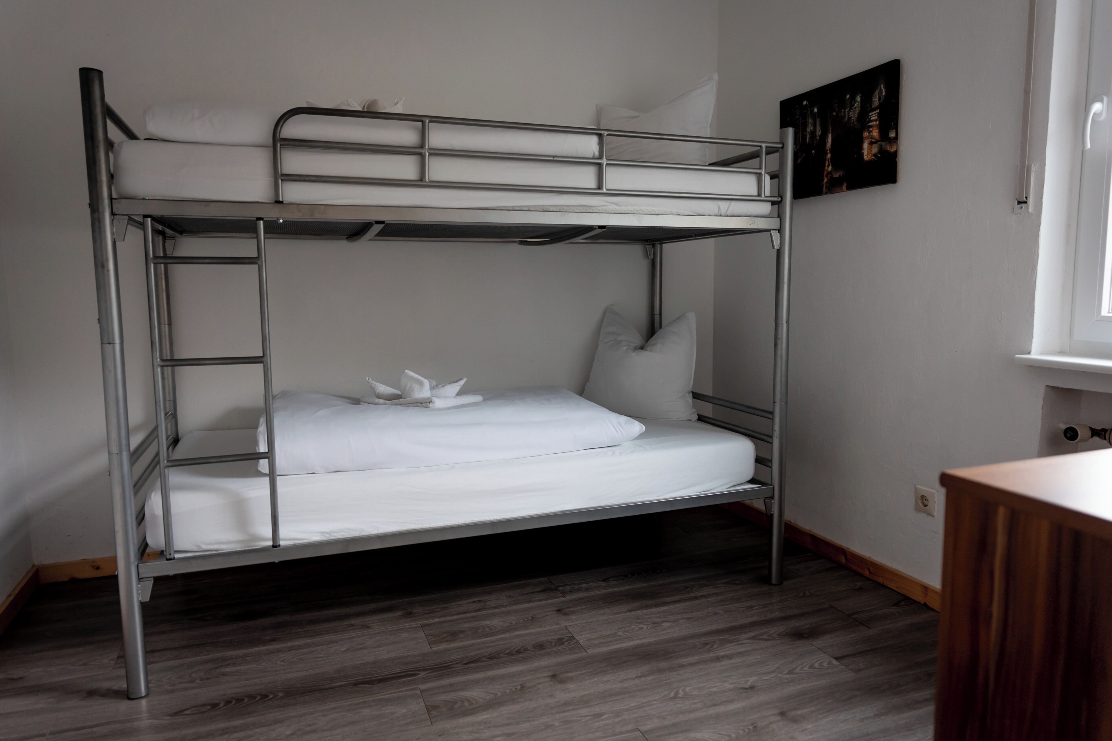 A bunk bed with white bedding, a wooden floor, a painting on the wall, and a window with white curtains.