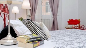 Ein ordentlich eingerichtetes Schlafzimmer mit Bett, Nachttischlampen, einem Nachttisch mit Büchern, einem Fenster mit Vorhängen und einem dekorativen Kopfteil.