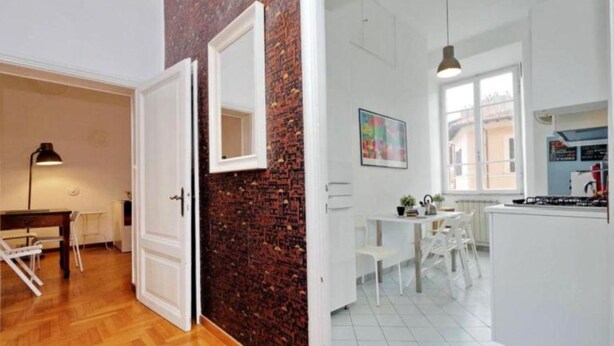 Ein Zimmer mit Holzboden, einer weißen Tür, einer Ziegelwand, einem weißen Tisch mit Stühlen sowie einer weißen Küchenzeile mit Herd und einer Pendelleuchte.
