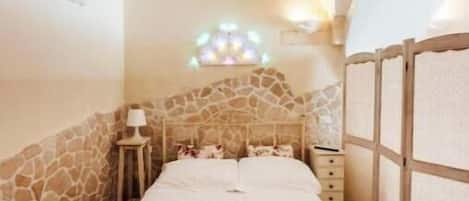 A narrow room with a stone wall, a bed with white linens, and a small table with a lamp.