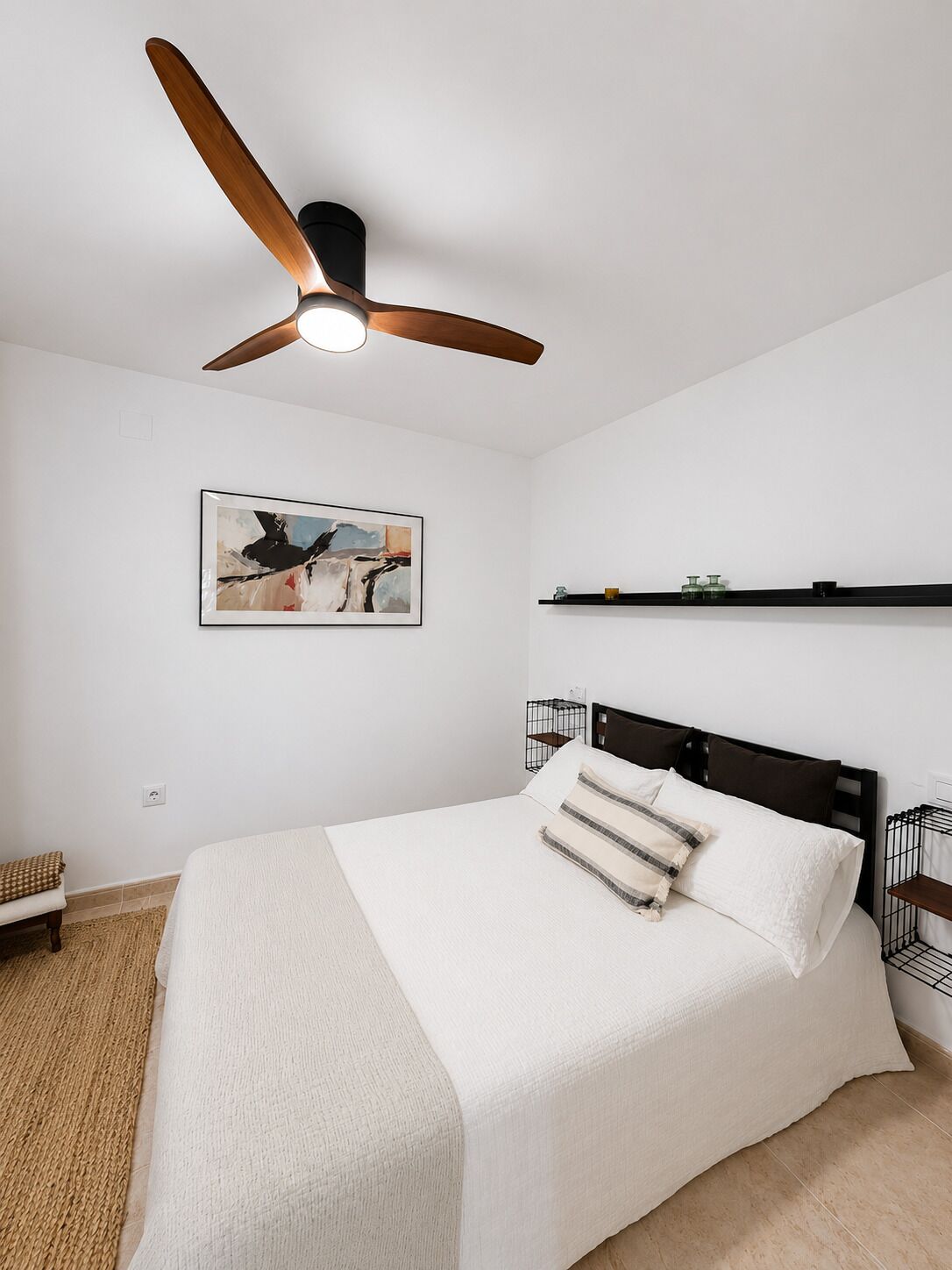 Un dormitorio con cama, ventilador de techo, estante en la pared y una obra de arte enmarco.
