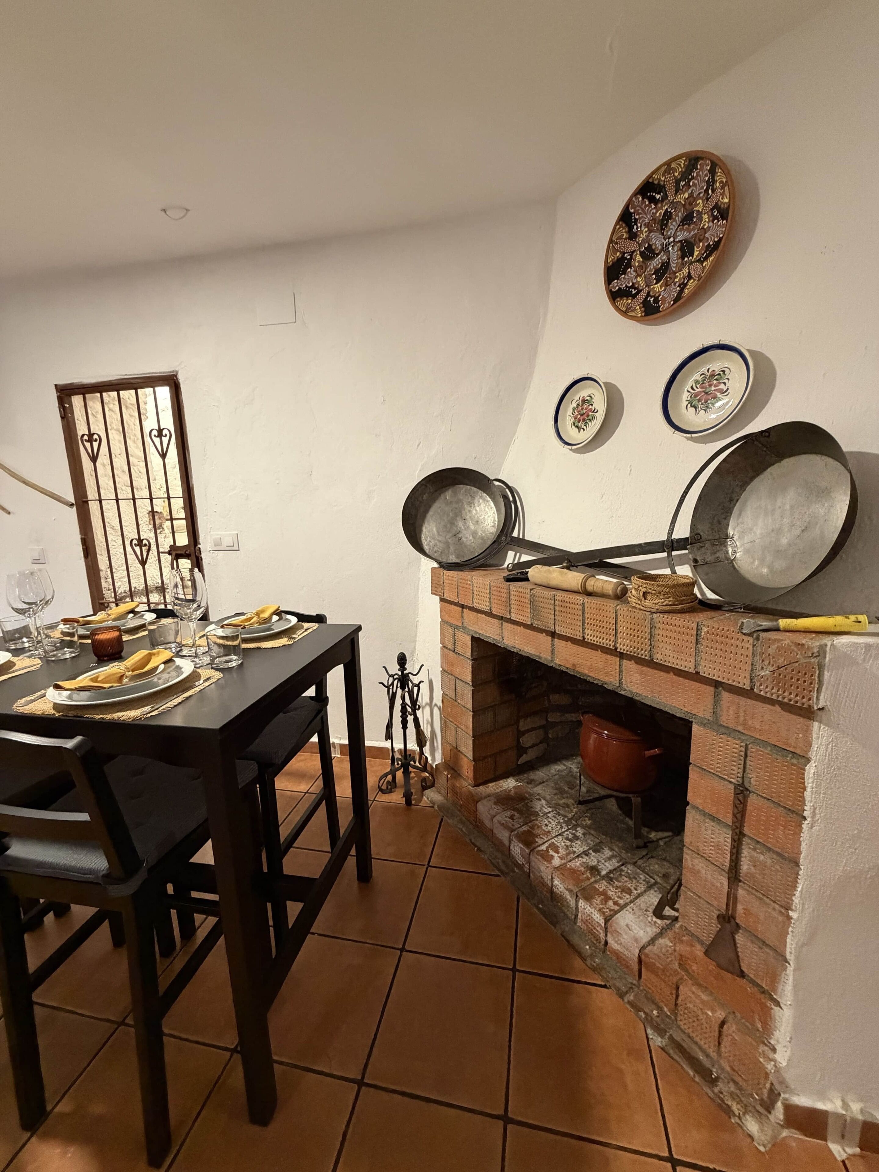 A dining area with a brick fireplace, a table set for two, and decorative plates on the wall.