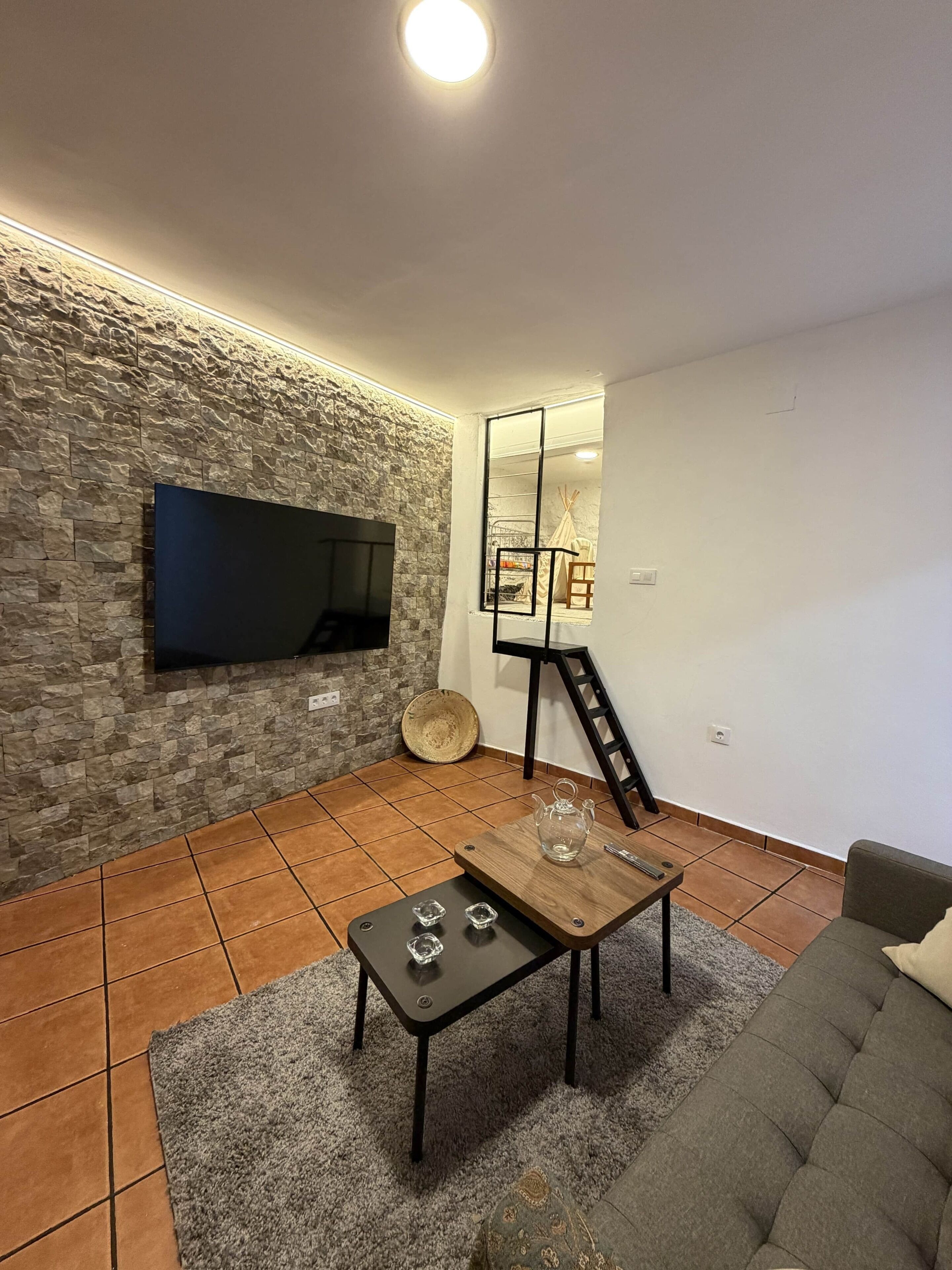 A living room with a stone wall, a flat-screen TV mounted on it, a wooden coffee table with a glass teapot, a grey sofa, and a staircase leading to a balcony.