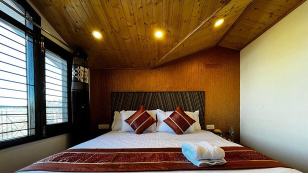 A hotel room with a wooden ceiling, a large bed with multiple pillows, a window with blinds, and a nightstand with a lamp.