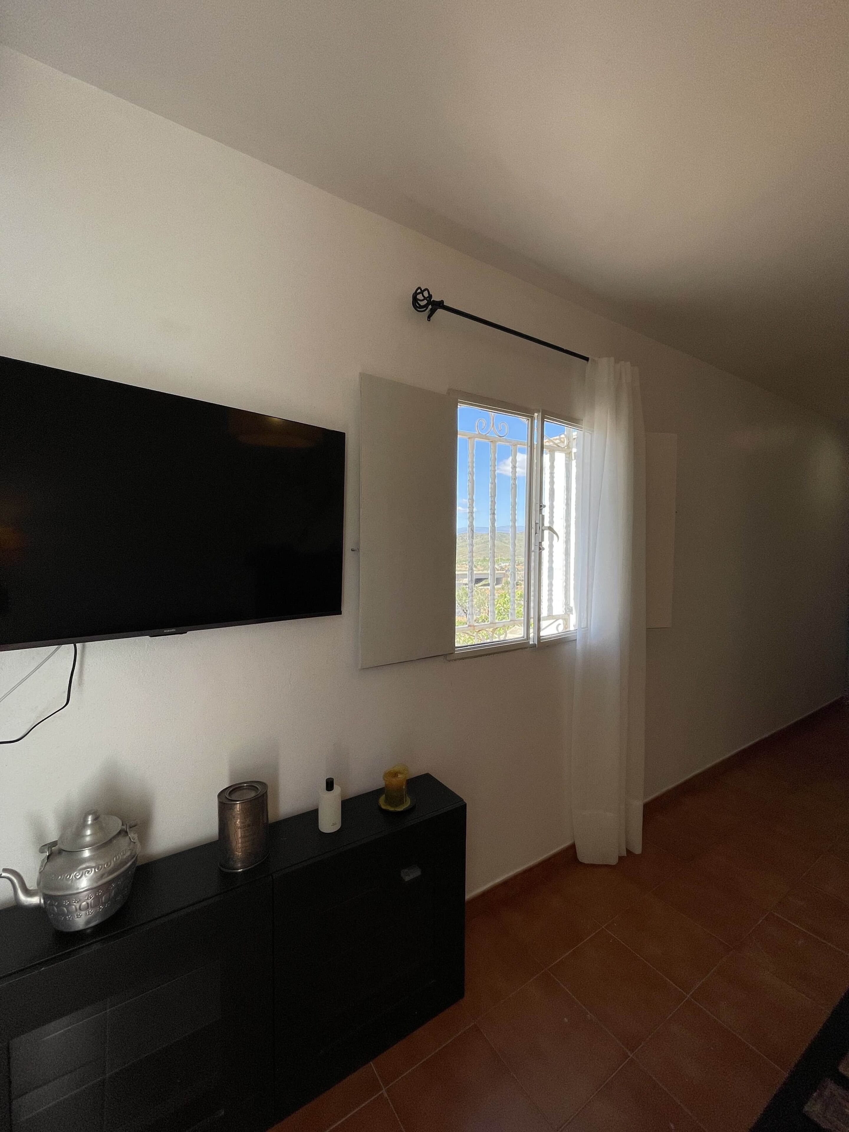 Una habitación con una televisión de pantalla plana montada en la pared, una ventana con cortinas blancas, un mueble negro con una tetera y una vela, y suelo de baldosas.