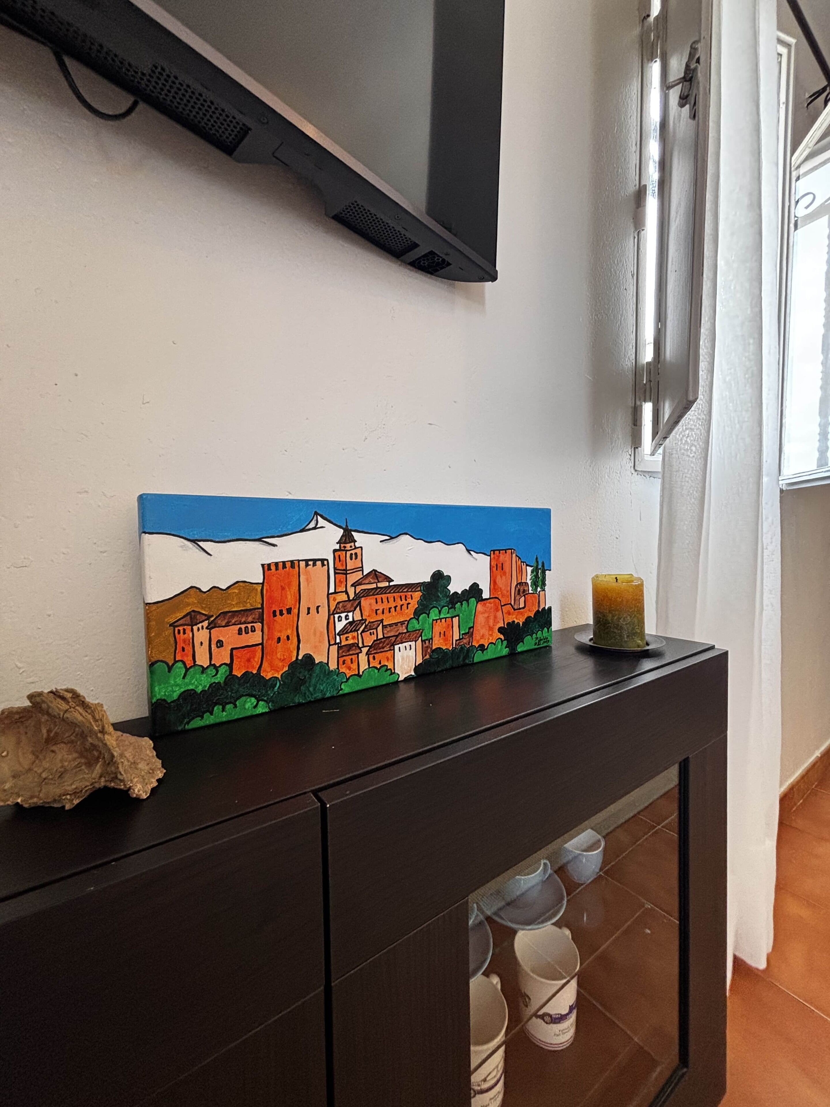Une peinture représentant une ville est accrochée sur une commode en bois foncé. Au-dessus, on aperçoit un téléviseur, et à droite, une fenêtre est ornée de rideaux.