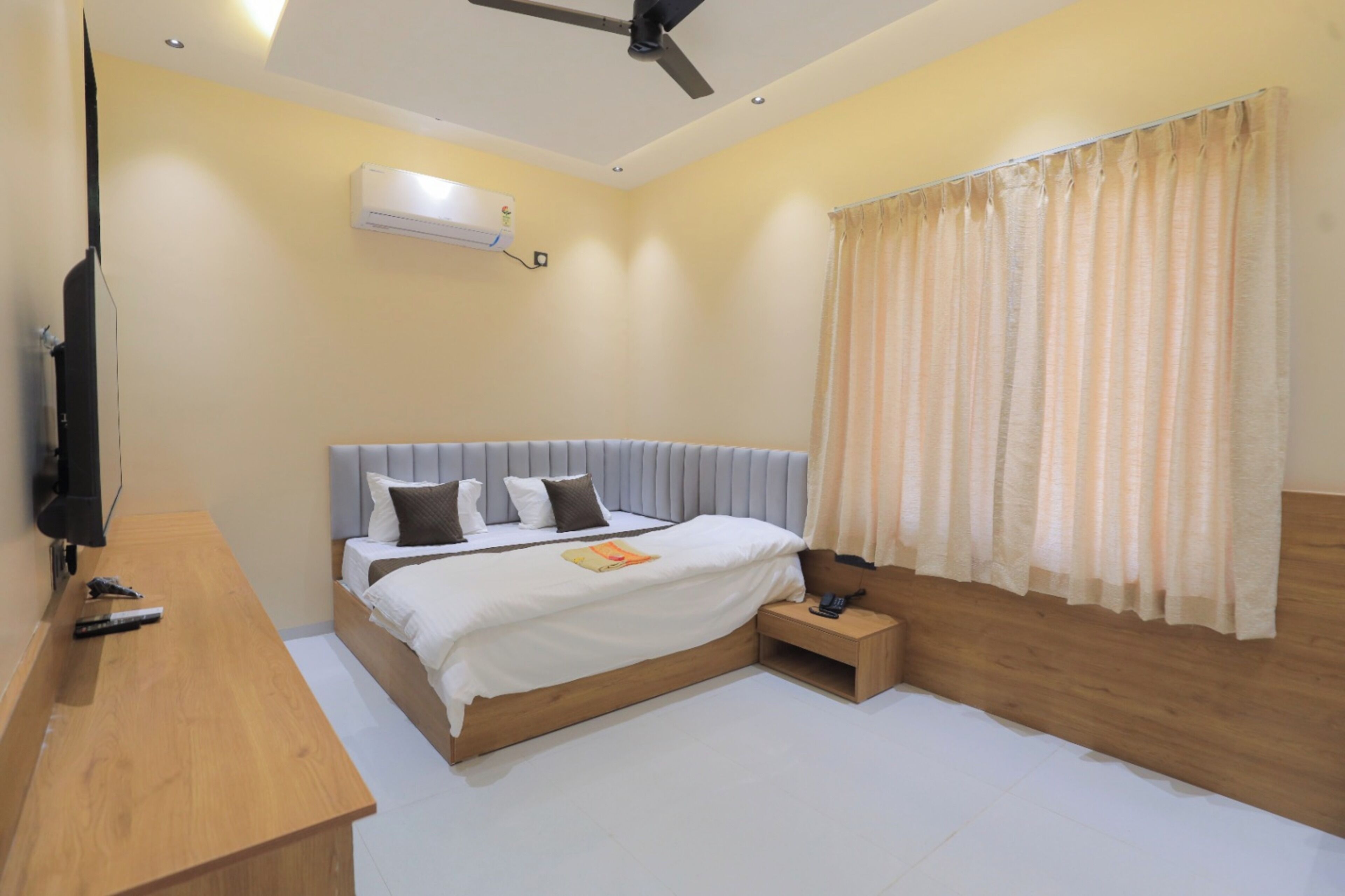 A bedroom with a bed, bedside table, nightstand, ceiling fan, air conditioning unit, and a window with curtains.