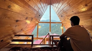 A wooden interior with a sloped ceiling, a view through a large window, and a person sitting on a bed. - The Woodshed (Mukteshwar)