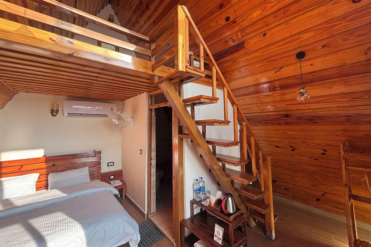 Interior d'una cabana de fusta amb un llit, una escala, un aparell d'aire condicionat i una taula petita amb objectes a sobre.
