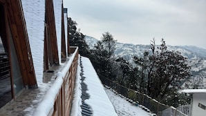 A snow-covered pathway leading to a building with a wooden railing and a view of snow-covered hills. - The Woodshed (Mukteshwar)