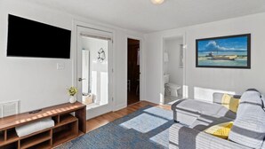 Una sala de estar con un sofá, un televisor montado en la pared, un cuadro y una puerta que da a otra habitación.