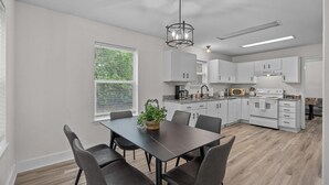 Una cocina moderna con armarios blancos, una mesa de comedor con sillas, una araña y una ventana con persianas.