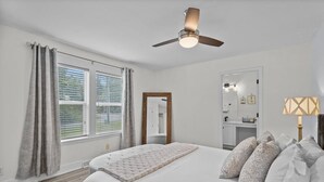 Un dormitorio con cama, ventilador de techo, ventana con cortinas, espejo y lámpara de noche.