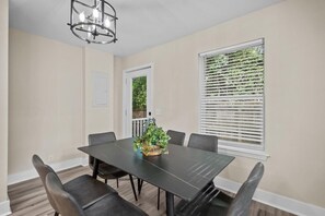 A dining area with a dark table, six chairs, a chandelier, a window with blinds, and a door.