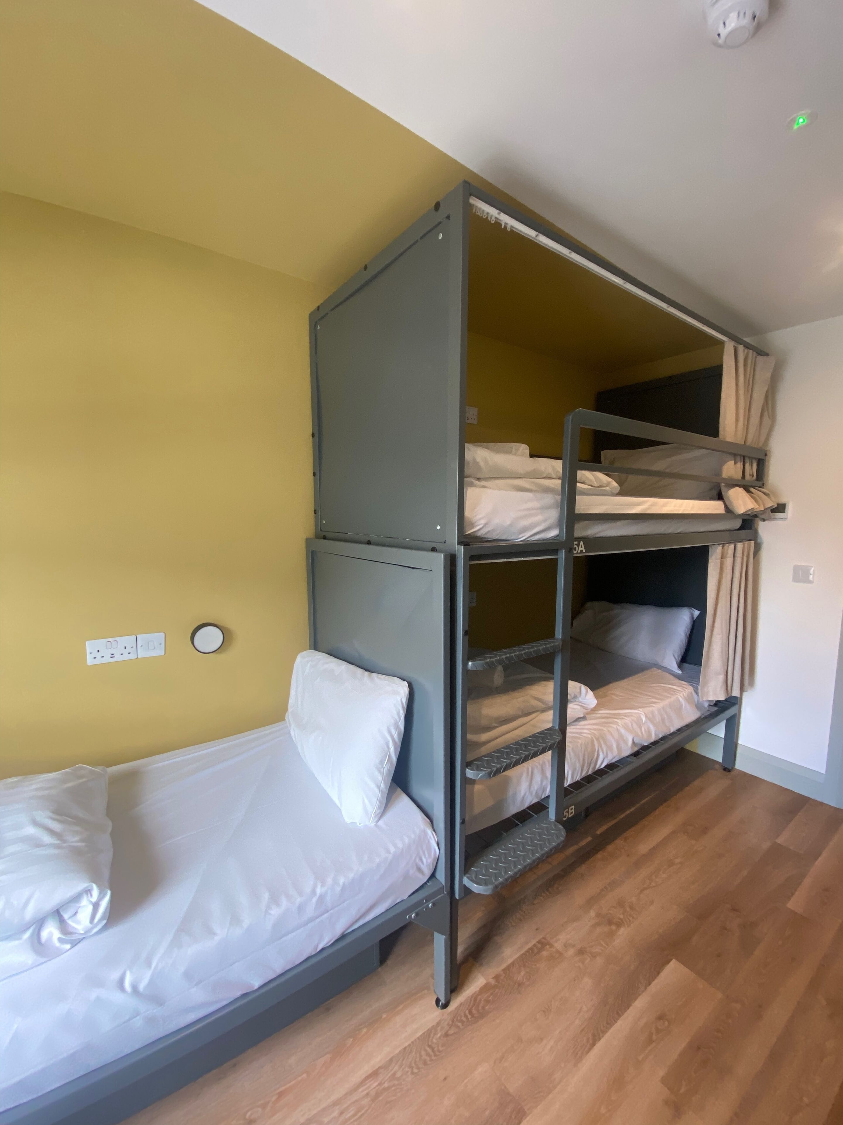 A bunk bed with two levels, a single bed on the left, and a wall socket and light switch on the wall.