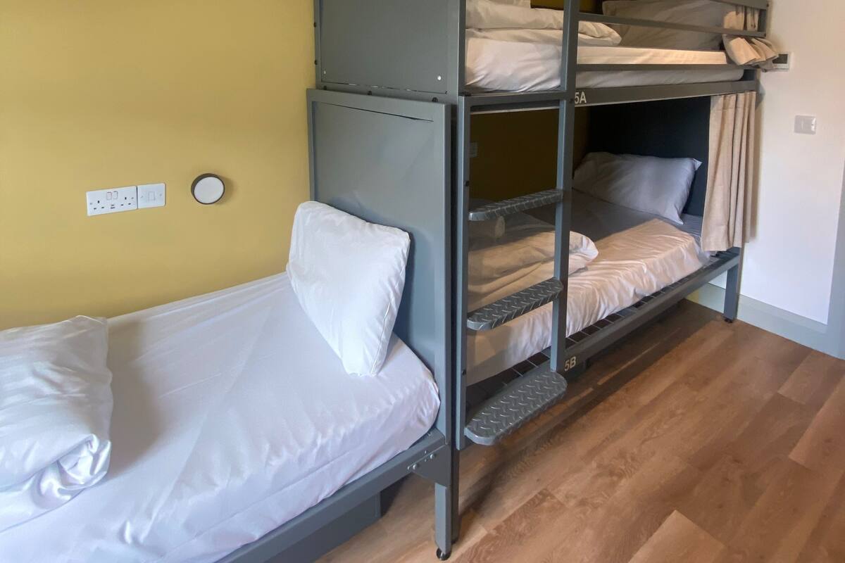 A bunk bed with two levels, a single bed on the left, and a wall socket and light switch on the wall.