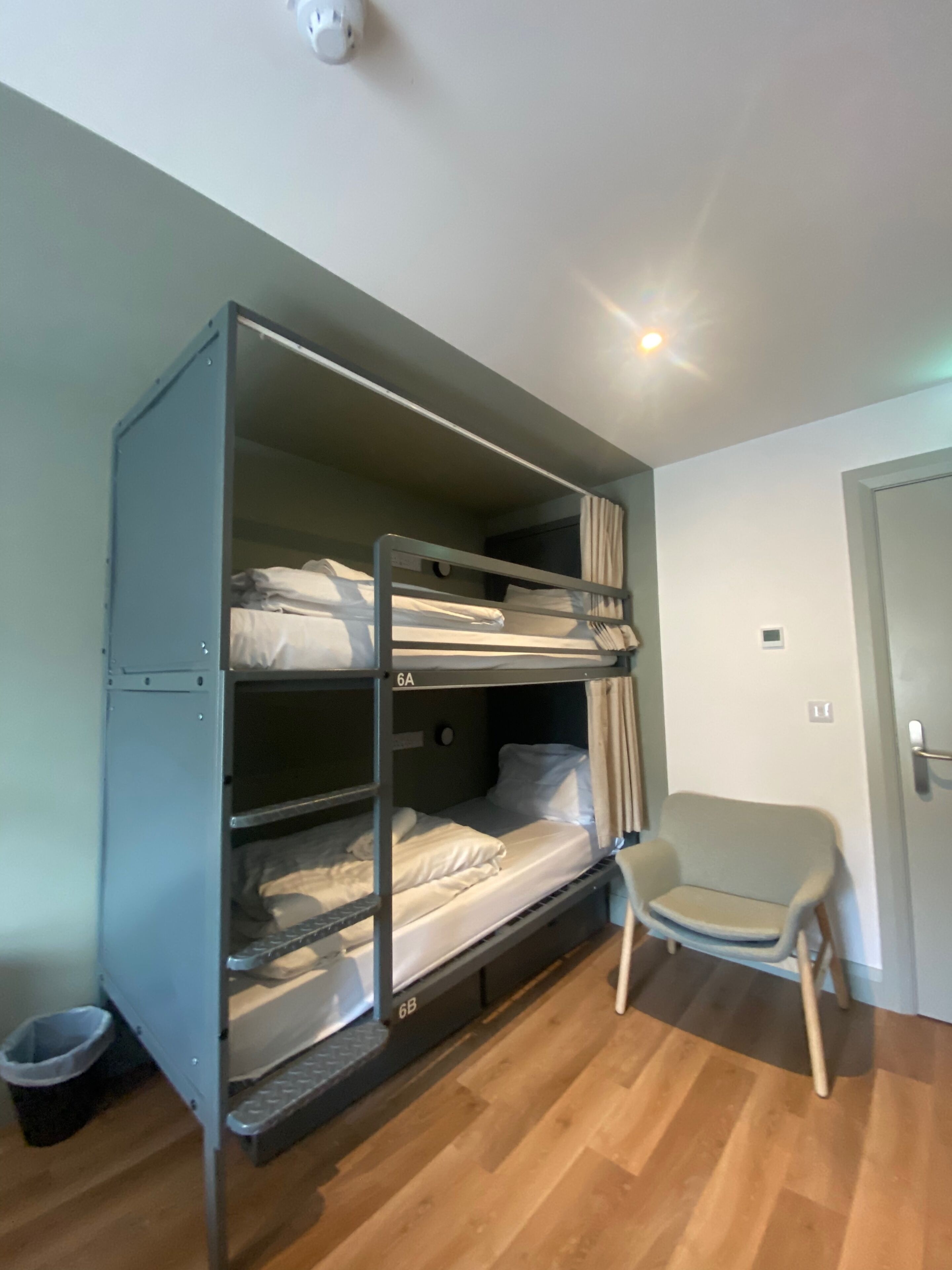 A bunk bed with two levels, a small chair, a trash bin, and a door.